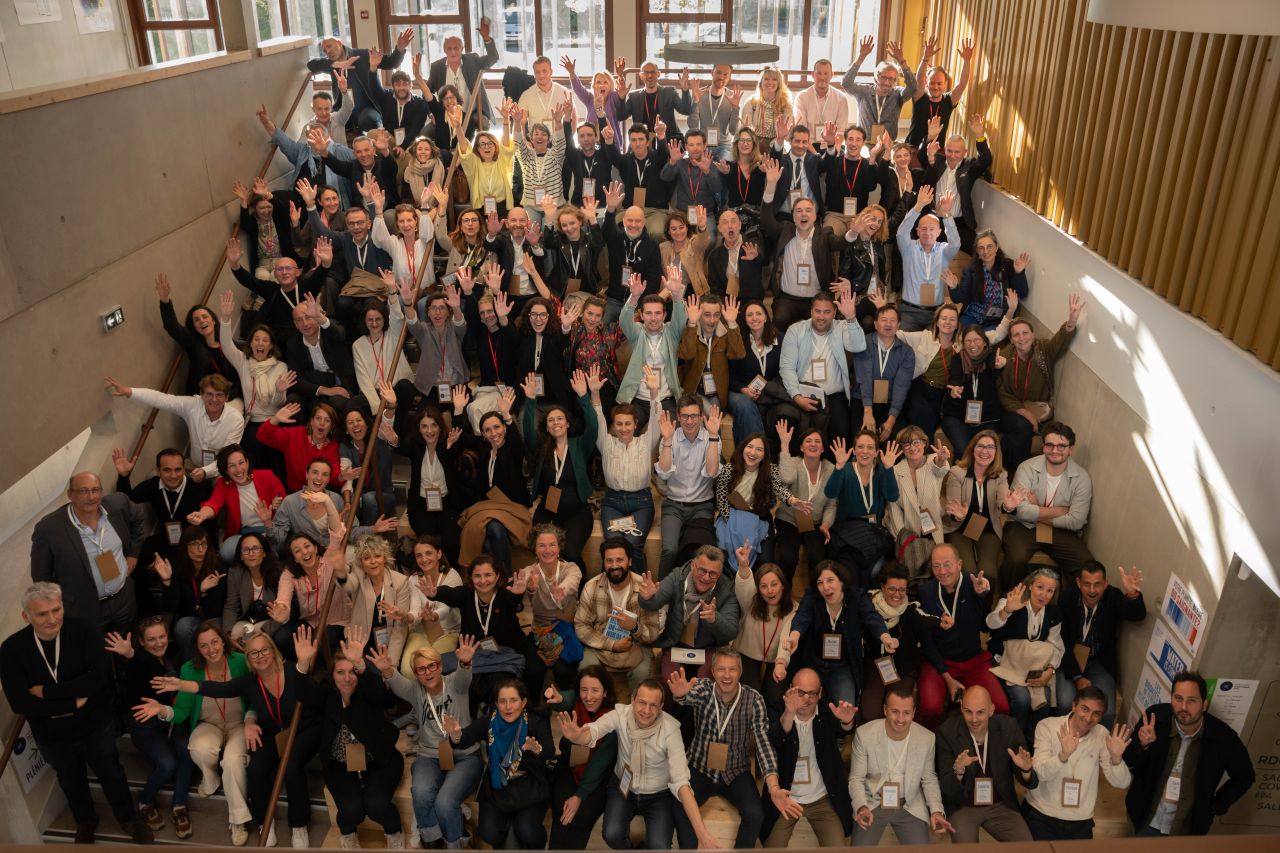 Groupe d’environ une centaine de personnes réunies sur un large escalier intérieur baigné de lumière naturelle. Les participants, portant des badges autour du cou, lèvent les mains vers l’objectif pour une photo de groupe.